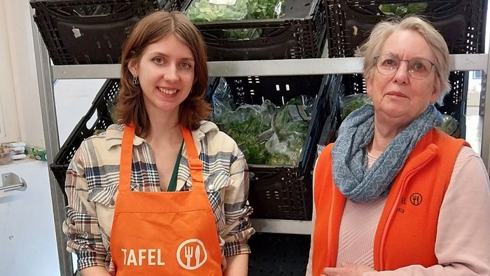 Zwei Frauen in orangefarbenen Schürzen und Westen stehen in einem Lager für Obst und Gemüse zwischen Kisten mit frischem Gemüse.