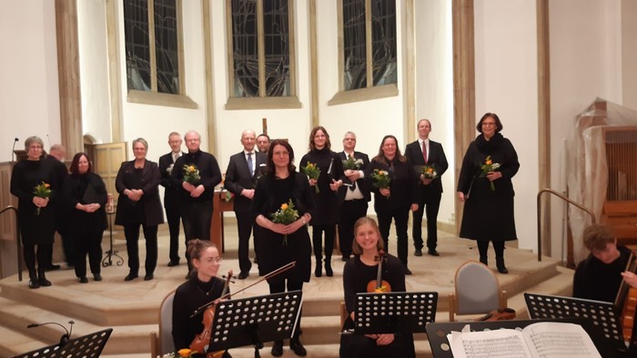 Ein Chor und Musiker treten in einer Kirche auf, halten Blumen und Notenblätter während einer offiziellen Veranstaltung.