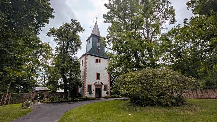 Das Bild zeigt eine kleine Kirche, die von Bäumen und Grünflächen umgeben ist. Der helle Bau hat einen schmalen Turm mit spitzem Dach, an dessen Spitze ein Kreuz angebracht ist. Rötliche Akzente an den Kanten heben sich vom hellen Mauerwerk ab. Vor der K
