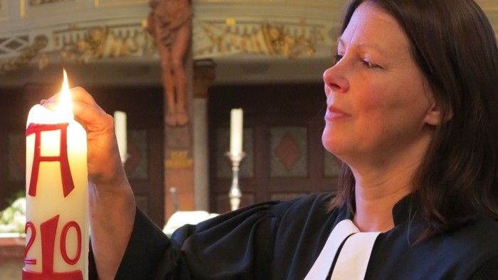 Eine Frau in einem Gewand hält eine brennende Kerze mit der Aufschrift „A210“ in einer Kirche.