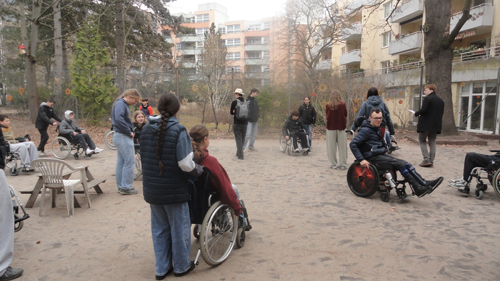 Menschen versammeln sich im Freien in einem Parkbereich, einige nutzen Rollstühle und andere helfen oder gehen in der Nähe.