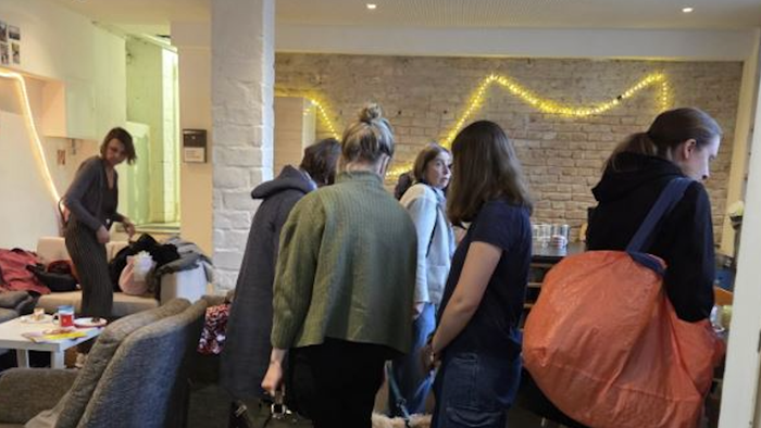 Eine Gruppe von Menschen versammelt sich drinnen bei festlicher Beleuchtung, plaudert und trägt Taschen in der Nähe eines gemütlichen Wohnbereichs.