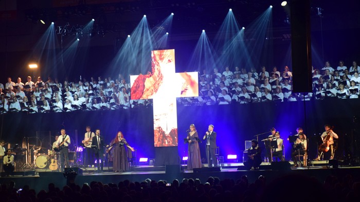 Großer Chor und Orchester treten auf der Bühne mit einer markanten Kreuzdarstellung während eines Abendkonzerts auf.