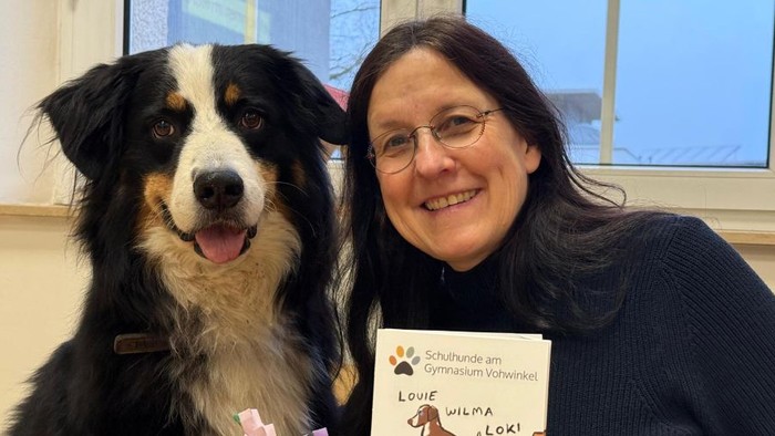 Eine Frau hält ein Buch mit ihrem lächelnden Hund und präsentiert eine Kindergeschichte über Schulhunde.