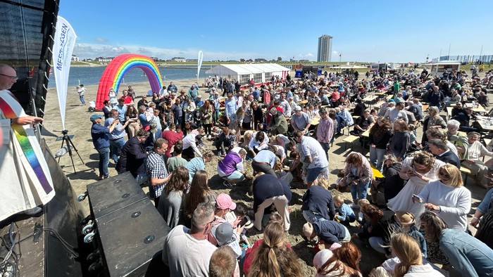 Eine große Menschenmenge versammelt sich im Freien zu einer lebhaften Veranstaltung am Wasser mit einem Regenbogenbanner und einer Bühne.
