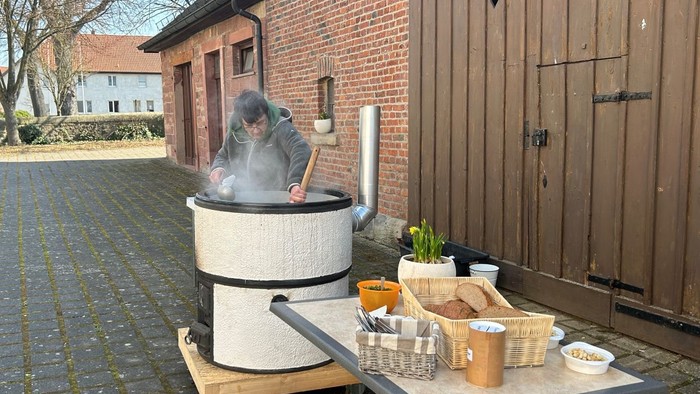 Person, die in einem großen Topf im Freien Essen zubereitet, in der Nähe eines rustikalen Ziegelgebäudes mit Vorräten auf einem Tisch.