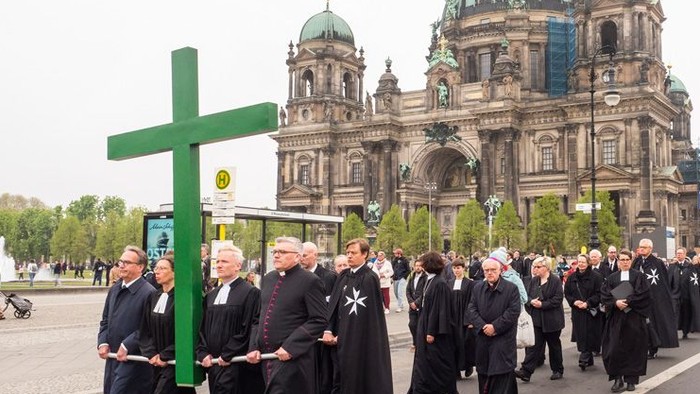 Pfarrerinnen und Pfarrer in schwarzen Talaren tragen ein sehr großes grünes Kreuz. Im Hintergrund ist der Berliner Dom zu sehen.
