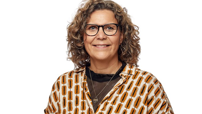 En glad person med krøllet hår, briller og en mønstret blazer, der smiler til kameraet.