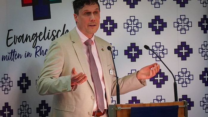 Ein Mann steht an einem Rednerpult und spricht in zwei Mikrofone. Er trägt ein helles Sakko, ein Hemd und eine Krawatte und gestikuliert mit geöffneten Händen, als würde er einen Punkt erläutern oder unterstreichen. Hinter ihm befindet sich eine Fotowand