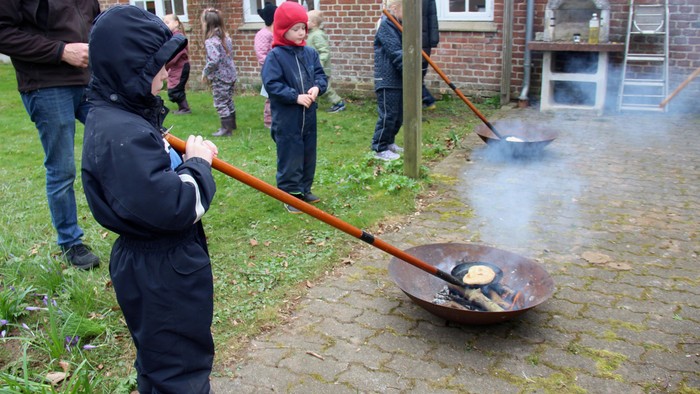 To bålfade med bål, og to børn, der holder en bålpande med brød på. I baggrunden børn, der kigger på