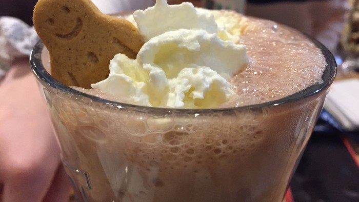 A glass mug filled with frothy hot chocolate topped with whipped cream and a smiling gingerbread cookie.