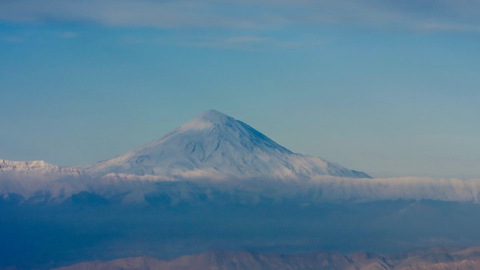 Majestätischer schneebedeckter Vulkan, der über wolkenverhangene Täler unter einem klaren blauen Himmel aufragt.