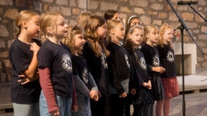 Eine Gruppe junger Kinder in einheitlichen Shirts, die auf einer Bühne mit Notenständern singen.