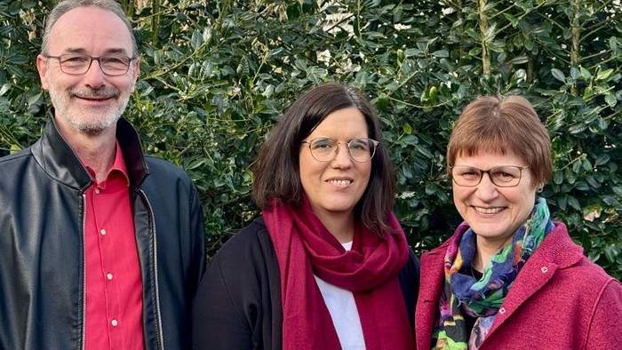 Drei lächelnde Erwachsene in Winterkleidung posieren gemeinsam vor einem grünen Heckenhintergrund.