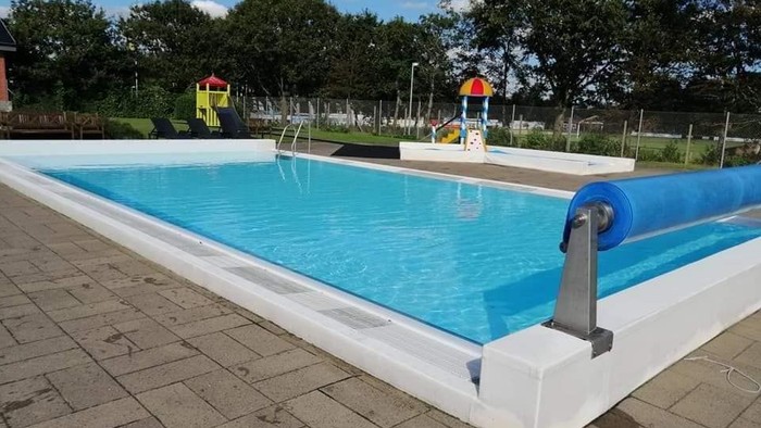 Et rektangulært svømmebasin med blå vand, omgivet af paver og med træer i baggrunden