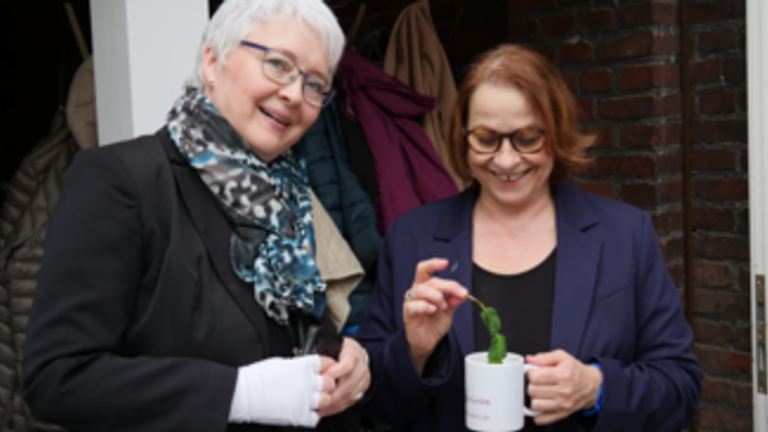 Zwei Frauen halten kleine Topfpflanzen und lächeln draußen in der Nähe eines Gebäudeeingangs.