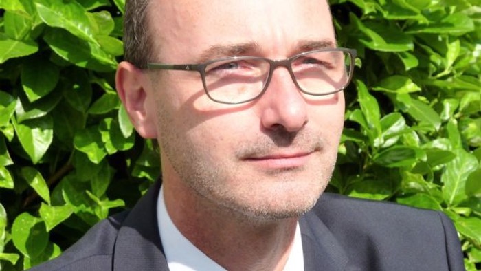 Ein Mann in einem dunklen Anzug und einer grünen Krawatte posiert vor üppigem grünem Blattwerk.