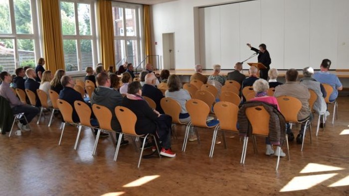 Eine Gruppe von Menschen hört einem Sprecher in einem hell erleuchteten Konferenzraum mit Holzbestuhlung aufmerksam zu.