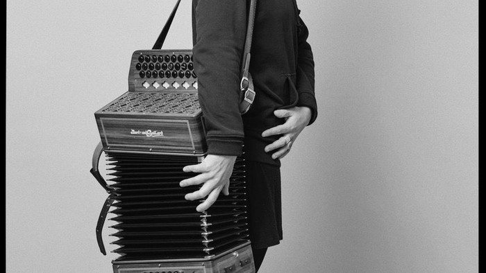 Frau mit Akkordeon, trägt eine Brille und Turnschuhe.