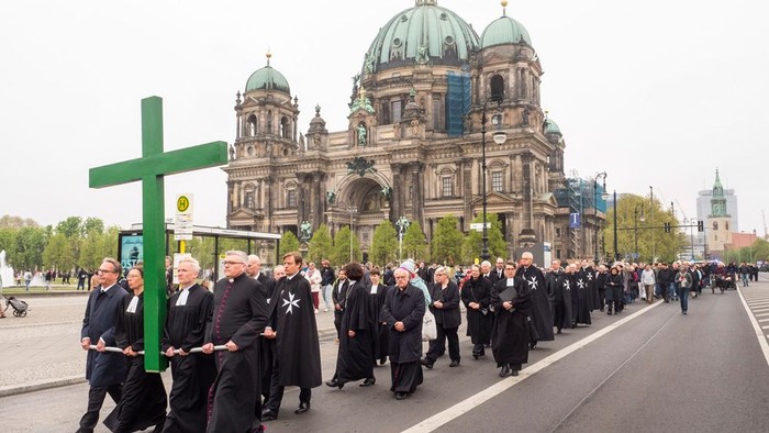 Pfarrerinnen und Pfarrer in schwarzen Talaren tragen ein sehr großes grünes Kreuz. Im Hintergrund ist der Berliner Dom zu sehen.