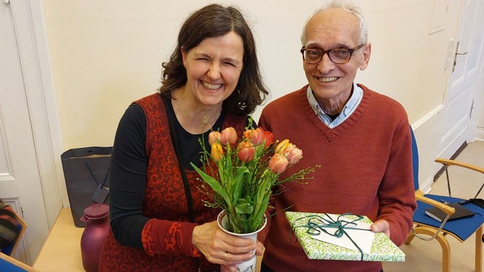 Eine lächelnde Frau und ein älterer Mann stehen nebeneinander in einem Raum, halten Blumen und ein Geschenk in den Händen und wirken festlich und glücklich.