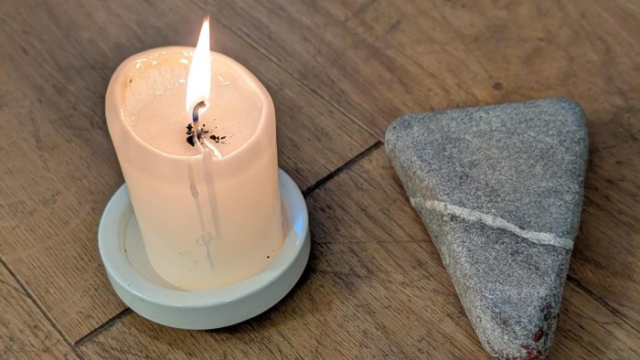 Eine brennende Kerze auf einem Untersetzer neben einem dreieckigen Stück rauen Steins auf einem Holzboden.