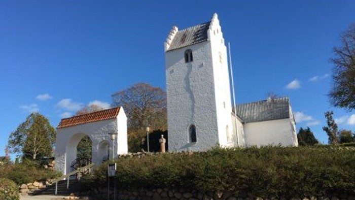 Et hvidt tårn og en lille kirke med rødt tag, omgivet af grønne buske og træer under en klar blå himmel