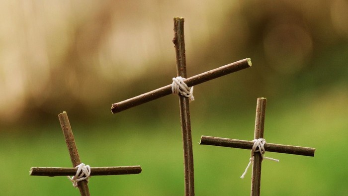 Drei einfache Holzkreuze, mit Schnur zusammengebunden, in einer grasbewachsenen Umgebung im Freien.