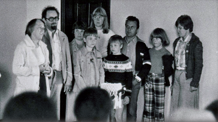 Mehrere Erwachsene und Kinder stehen gemeinsam auf einer Bühne; im Vordergrund sind die Köpfe von Zuschauenden zu sehen. Schwarzweiss-Bild