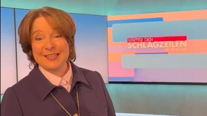 Eine Frau in geistlicher Kleidung lächelt in einem farbenfrohen Nachrichtenstudio und diskutiert die Schlagzeilen hinter ihr.