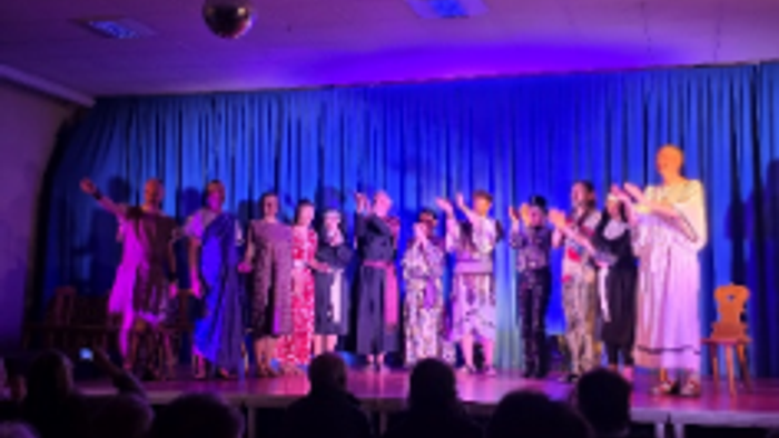 Eine Gruppe von Darstellern auf der Bühne in bunten Kostümen, die unter lila beleuchteten Vorhängen singen oder tanzen.