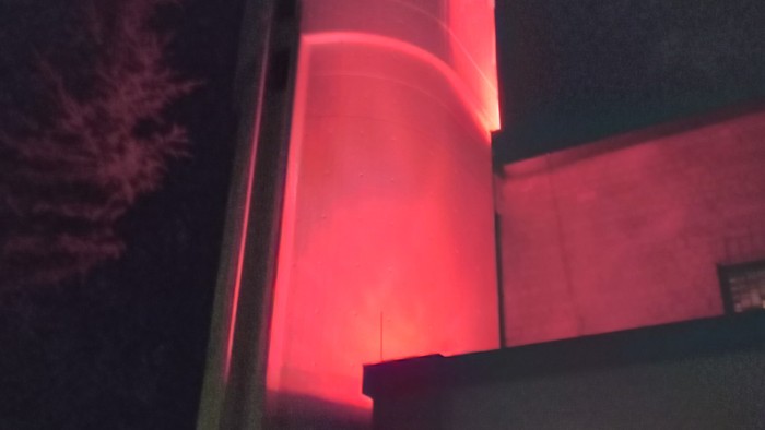 Eine hohe zylindrische Struktur, die nachts mit hellem rotem Licht beleuchtet wird, in der Nähe eines Gebäudeeingangs.