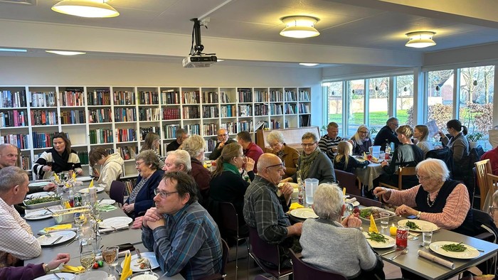En gruppe ældre personer spiser sammen i en lokal med mange bøger på vægge.