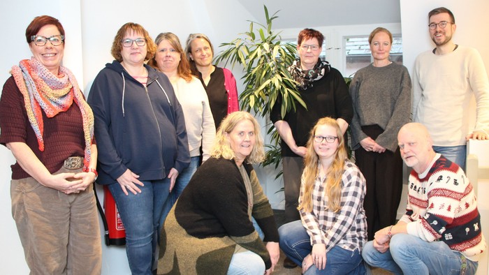 Eine fröhliche Gruppe von neun Personen, die drinnen gemeinsam posieren, mit einer Pflanze im Hintergrund.