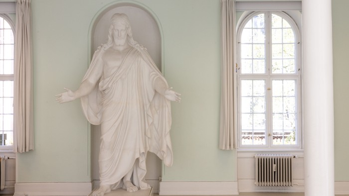 En klassisk marmorstatue af en klædt figur står yndefuldt i et lysegrønt rum med buede vinduer.