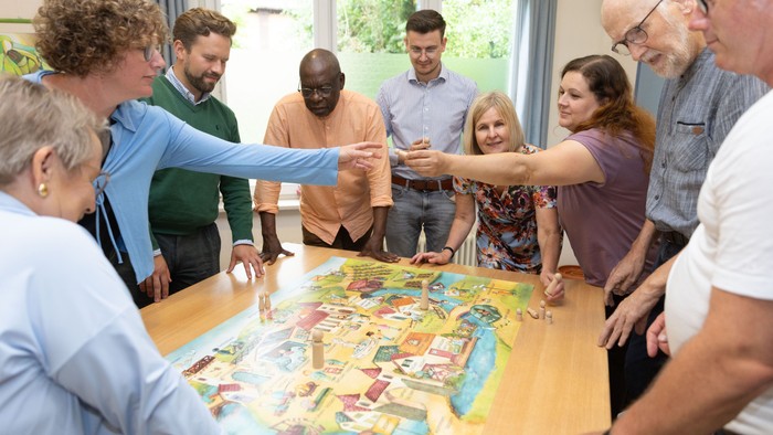 Eine vielfältige Gruppe spielt gemeinsam ein Brettspiel an einem großen Tisch in einem gemütlichen Raum.