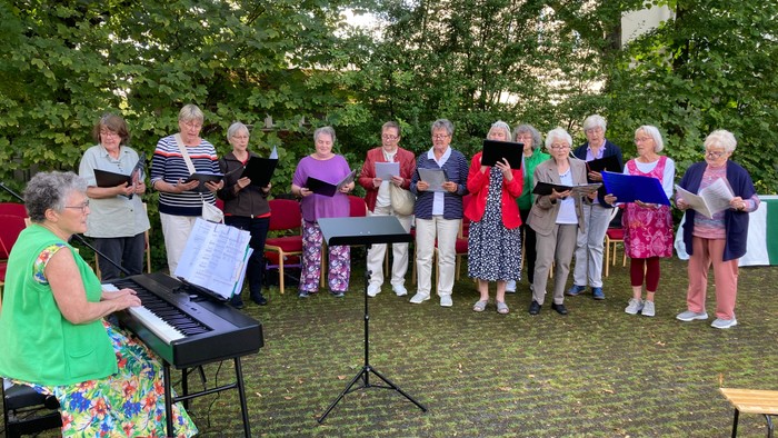 Eine Gruppe von Menschen, die im Freien singen, begleitet von einem Pianisten in einer Parklandschaft.