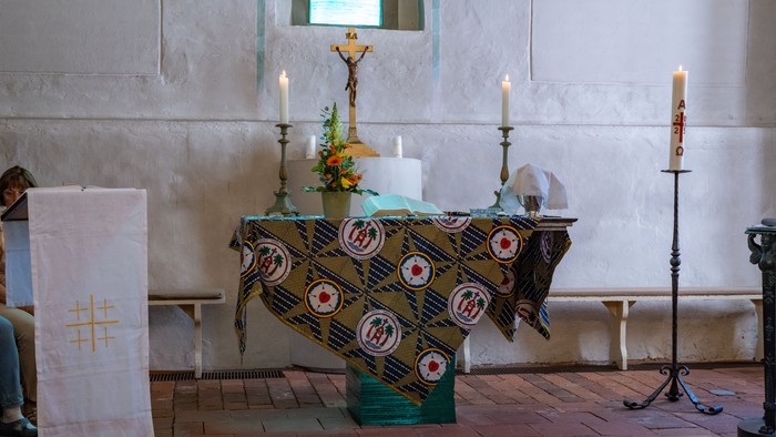 Altarraum der Dorfkirche Alt-Staaken mit weiß verputzter Wand, Kreuz, zwei brennenden Kerzen und einem Altar, der mit einem farbigen Partnerschaftstuch geschmückt ist. Links steht ein weißes Lesepult, rechts eine Osterkerze; der Raum wirkt schlicht, hell 