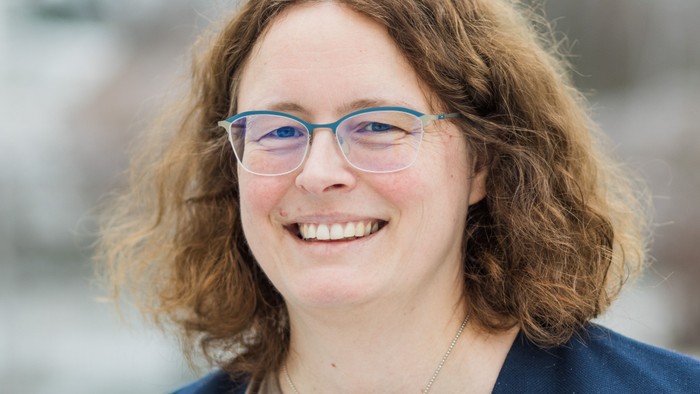 Eine fröhliche Person mit lockigem Haar, die eine Brille und einen blauen Blazer trägt, lächelt im Freien.