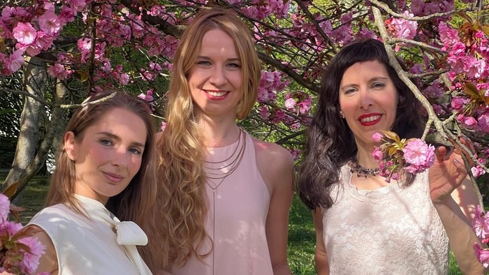 Drei Frauen in eleganter Frühlingskleidung posieren gemeinsam unter blühenden rosa Bäumen.