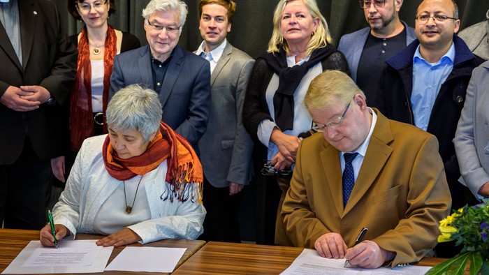 Zwei Personen unterschreiben Vertrag, umgeben von mehreren Beobachtern.