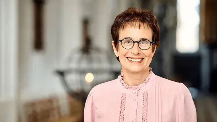 En glad kvinde i en lyserød bluse smiler varmt indendørs med dekorativ belysning i baggrunden.