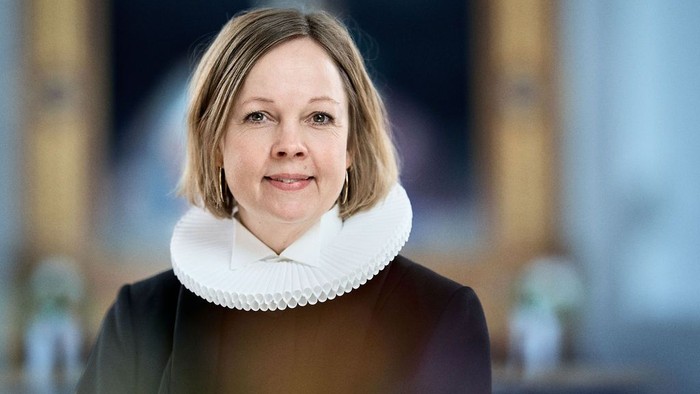 En person iført en mørk frakke med et hvidt højhalset tørklæde står indendørs nær en storslået arkitektonisk baggrund.