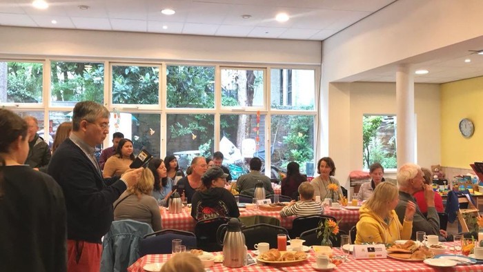 Eine Gruppe von Menschen sitzt an Tischen und isst in einem hellen Raum mit großen Fenstern.