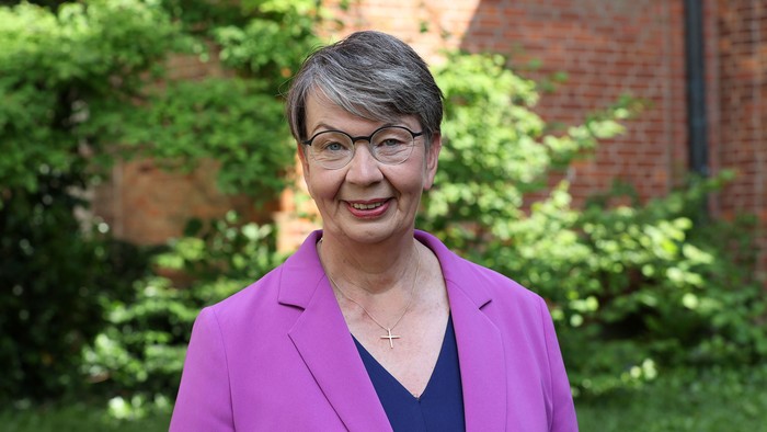 Eine Frau mit Brille und lila Blazer steht vor einem Ziegelgebäude und grünen Büschen.