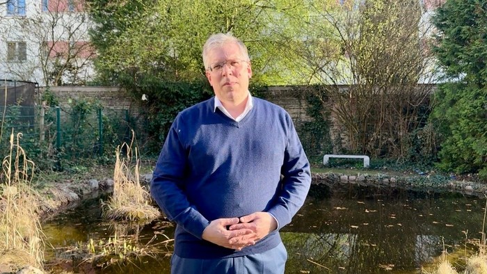 Ein Mann steht an einem Teich in einer ruhigen Umgebung im Freien mit Grünflächen und einer Backsteinmauer im Hintergrund.