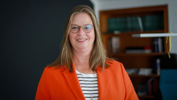 Eine Frau mit offenen Haaren, Brille und einem orangenen Jacket in einem Büro