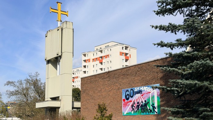 Moderne Kirche mit Kreuz auf dem Dach, angrenzend an eine Ziegelmauer mit farbenfrohem Kunstwerk zum 60-jährigen Jubiläum.