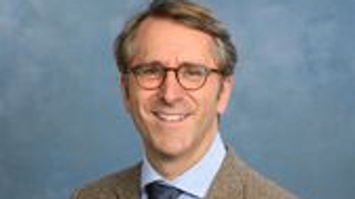 Professioneller Mann mit Brille, Tweedjacke und blauer Krawatte, der in die Kamera lächelt.