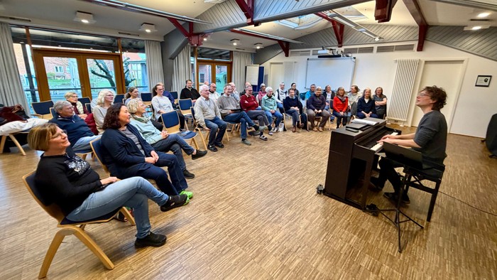 Eine Gruppe versammelt sich in einem geräumigen Raum, um einem Sprecher zuzuhören, der Klavier spielt und vorträgt.